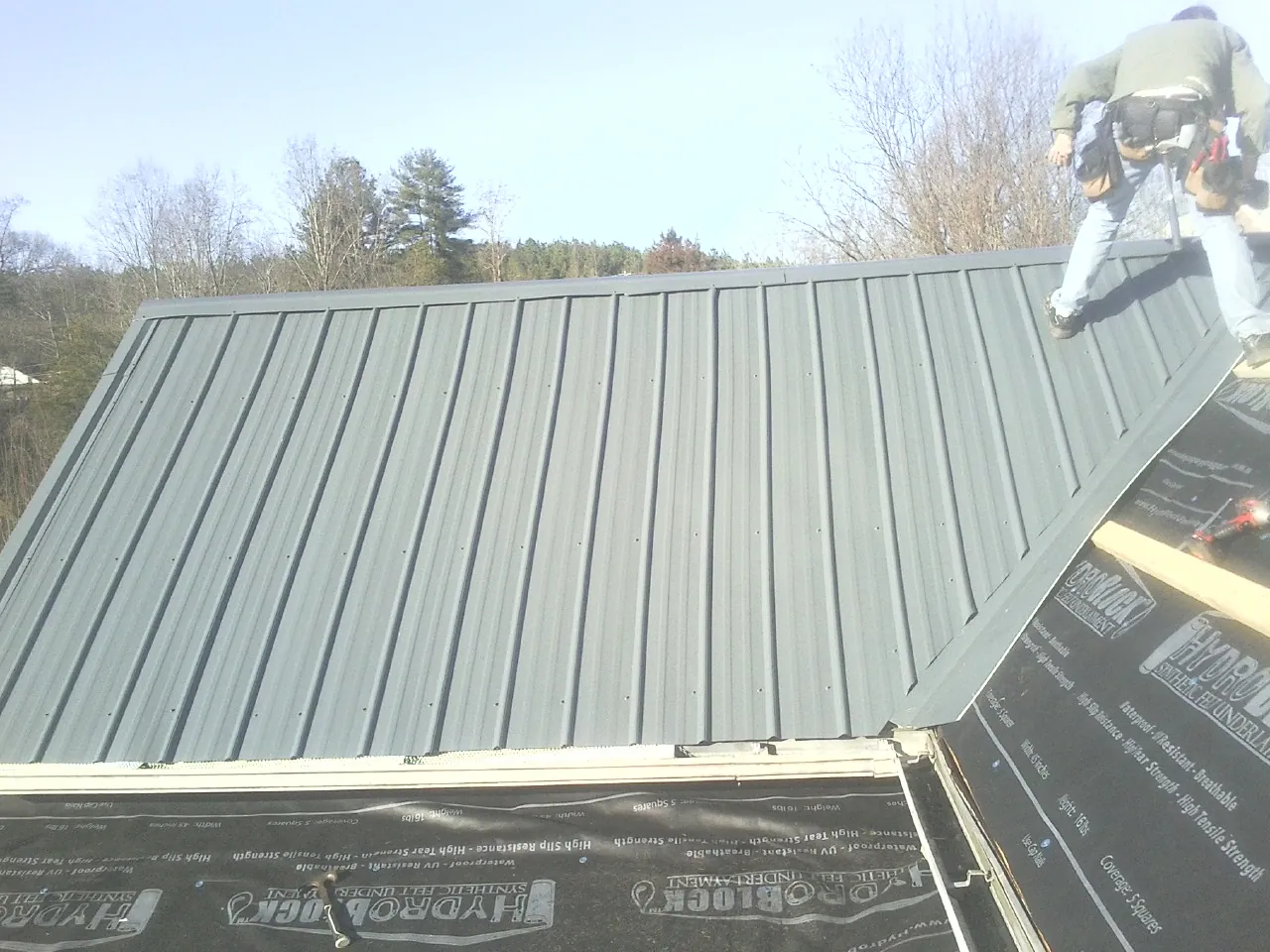 Crew installing dark green standing seam panels over synthetic underlayment in East Berne