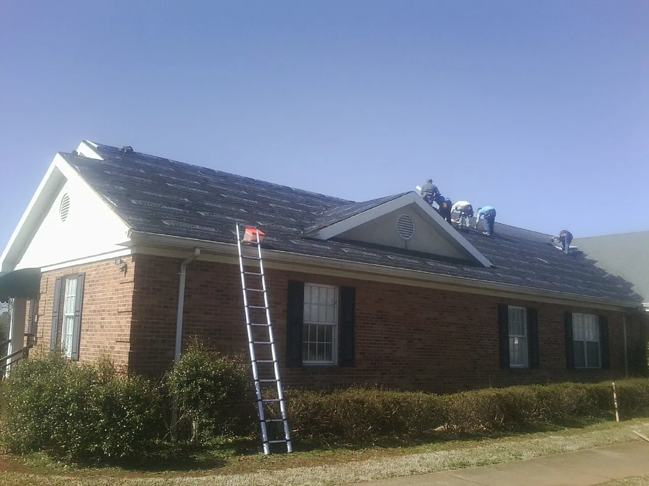 Crew tearing shingles off a brick ranch roof with a ladder set — Metal Roof Repair work by our crew in East Berne
