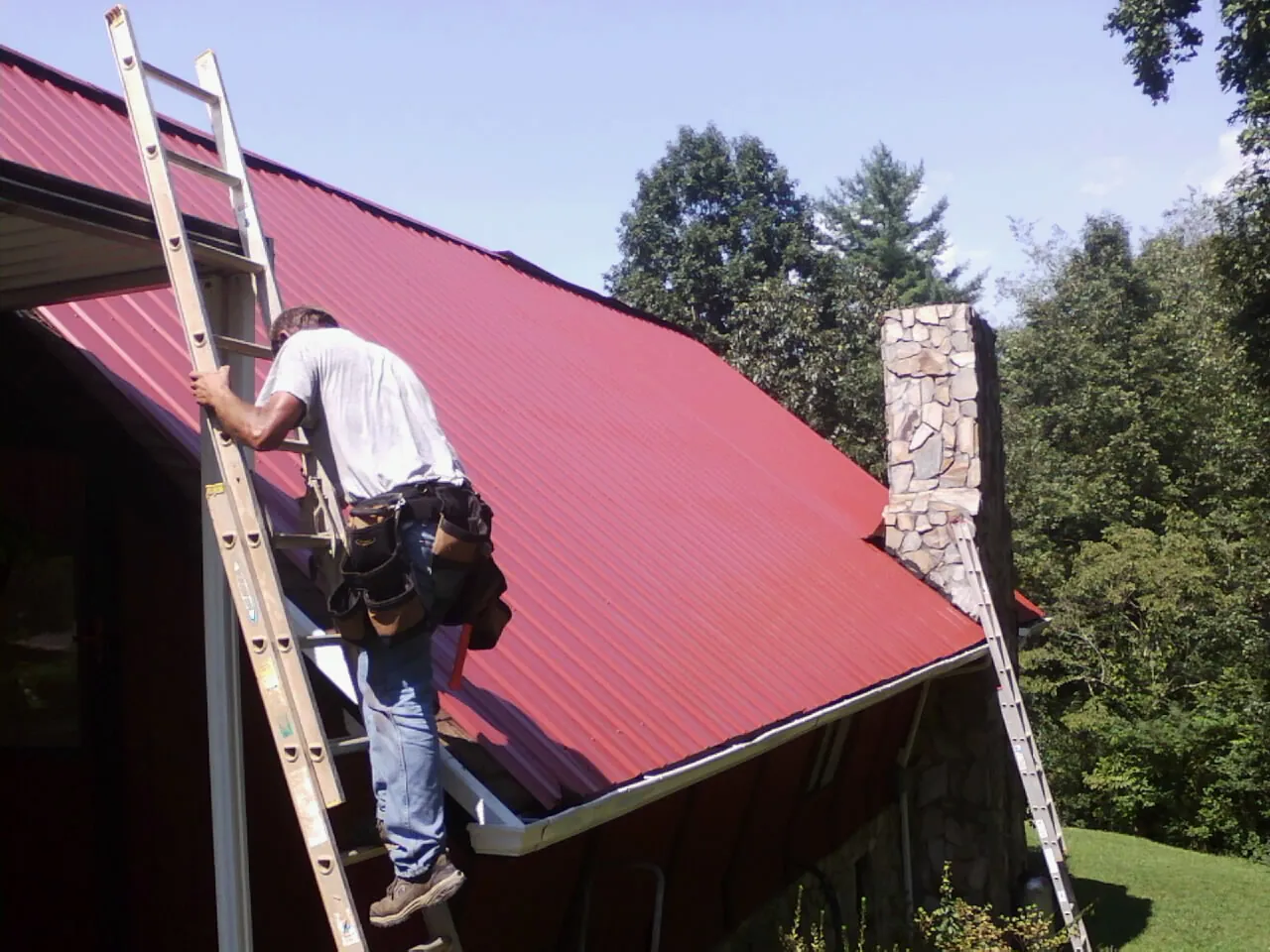 Roofer on a ladder beside a red metal roof and stone chimney — Soffit & Fascia Repair by Roofing Brothers in East Berne